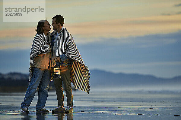 Affectionate couple wrapped in shawl holding lantern at ocean beach