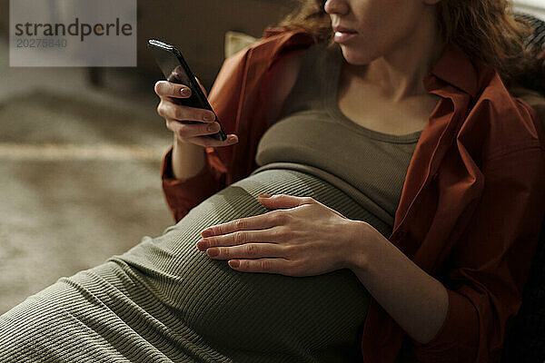 Pregnant woman touching belly and using mobile phone at home
