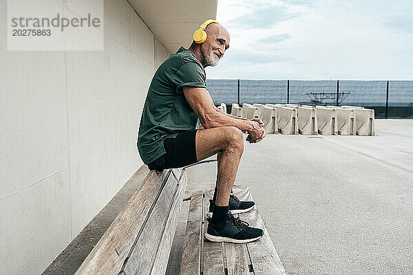 Lächelnder älterer Mann  der auf einer Bank sitzt und Musik über drahtlose Kopfhörer hört