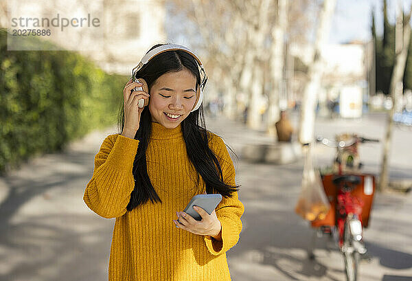 Lächelnde Frau trägt kabellose Kopfhörer und benutzt ein Smartphone