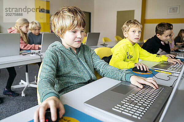 Blonder Junge benutzt Laptop und lernt Computerprogrammierung am Schreibtisch im Klassenzimmer