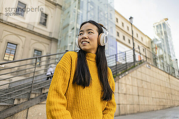 Lächelnde junge Frau mit kabellosen Kopfhörern in der Nähe eines Gebäudes