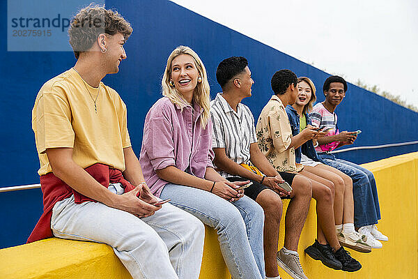Fröhliche Freunde in farbenfroher Kleidung sitzen an einer gelben Wand und halten Smartphones in der Hand