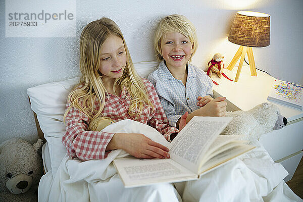 Happy siblings reading book sitting on bed at home