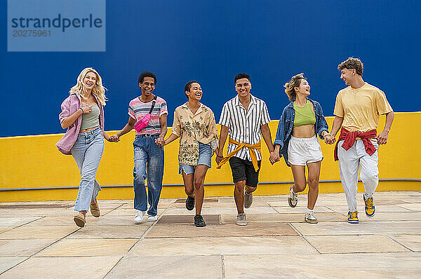 Multiethnische Gruppe junger Freunde  die Händchen haltend vor einer gelben Wand laufen