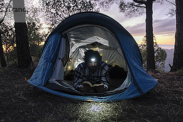 Mann trägt Stirnlampe und liest Buch im Zelt