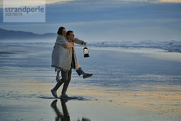 Junger Mann huckepack  Frau mit Laterne am Meeresstrand bei Sonnenuntergang