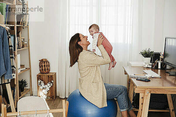 Mutter holt kleines Mädchen ab  das im Heimbüro auf einem Fitnessball sitzt