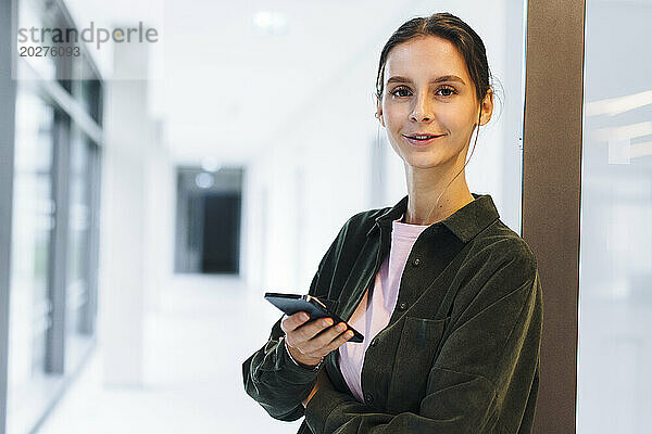 Lächelnde Geschäftsfrau steht mit Smartphone im Büroflur