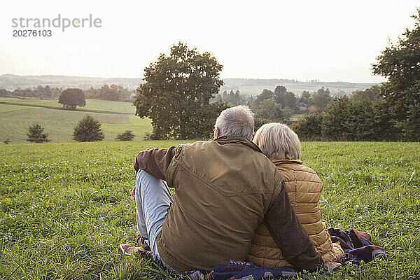 Rückansicht eines älteren Paares  das auf einer Wiese sitzt und den Sonnenuntergang genießt
