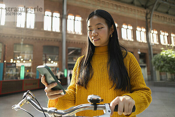 Lächelnde Frau mit Fahrrad und Smartphone am Bahnhof