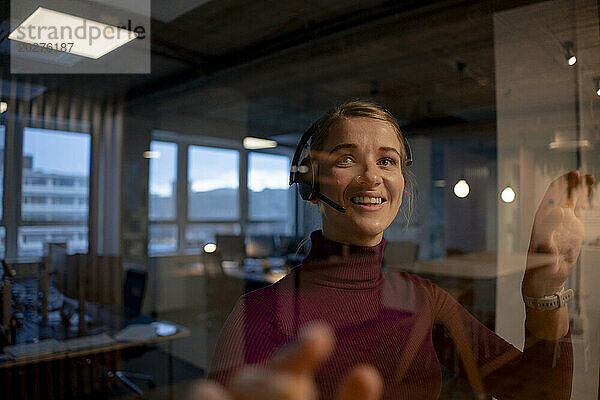 Zufriedener Kundendienstmitarbeiter mit Headset in der Nähe der Glaswand im Büro