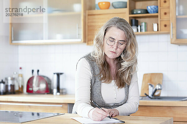 Ernsthafte reife Frau schreibt zu Hause in der Küche auf Papier