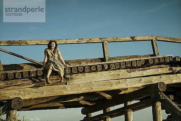 Sorglose Frau sitzt auf Holzbrücke unter blauem Himmel