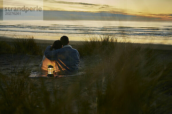 Junger Mann mit dem Arm um seine Freundin  der bei Sonnenuntergang am Strand sitzt