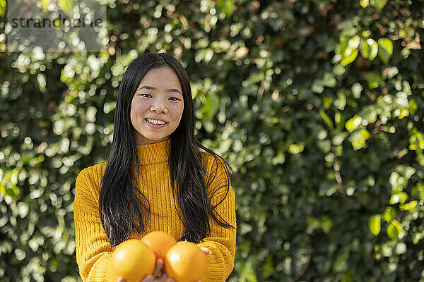 Lächelnde junge Frau hält Orangen vor Pflanzen