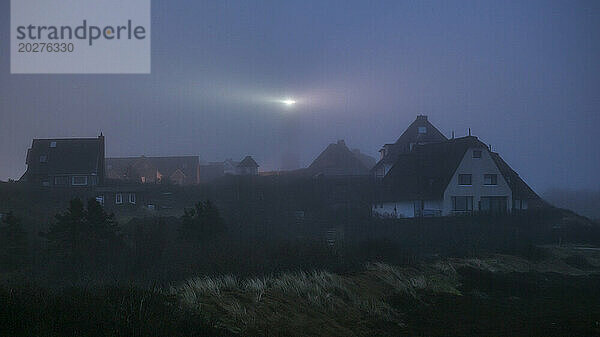 Deutschland  Schleswig-Holstein  Hörnum  Dorfhäuser im nebligen Morgengrauen mit leuchtendem Leuchtturm im Hintergrund