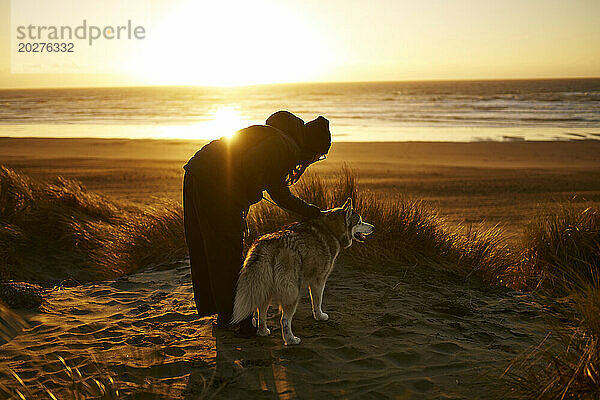 Junge Frau streichelt Hund am Strand bei Sonnenuntergang