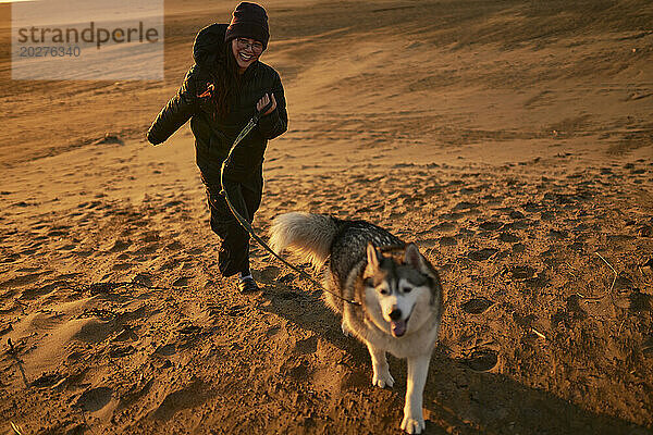 Cheerful woman running with dog on sand at sunset