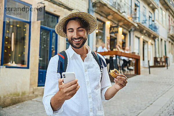 Glücklicher junger Mann hält traditionelles portugiesisches Dessert in der Hand und benutzt Smartphone