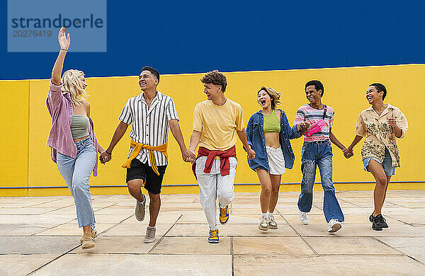 Multiethnische Gruppe junger Freunde  die Händchen haltend vor einer gelben Wand laufen