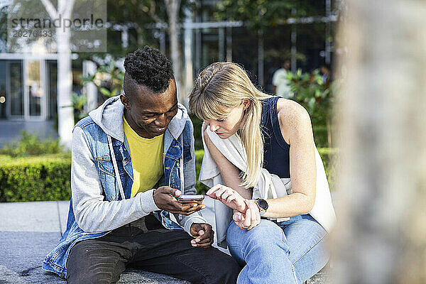 Neugierige  gemischtrassige Freunde  die auf einer Bank sitzen und ihr Smartphone teilen