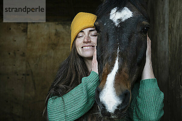 Smiling woman with eyes closed petting piebald horse in stable