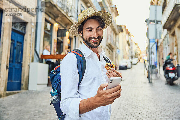 Lächelnder junger Mann benutzt Smartphone und hält traditionelles Dessert in der Hand