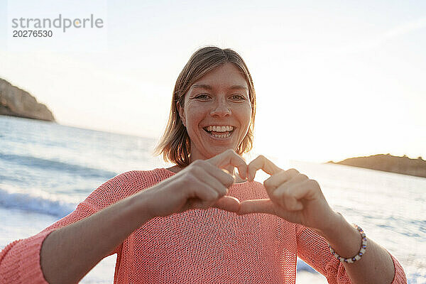 Carefree woman gesturing heart shape at sunset beach