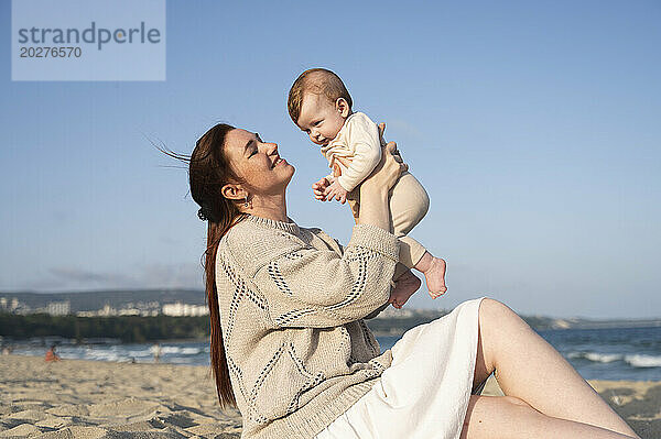 Lächelnde Frau sitzt auf Sand und spielt mit ihrem kleinen Mädchen am Strand