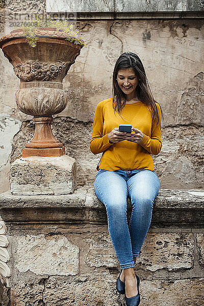 Lächelnde Frau benutzt Smartphone in der Nähe eines Terrakotta-Blumentopfs vor der Wand