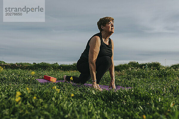 Aktive Seniorin praktiziert Yoga auf einer Trainingsmatte inmitten von Pflanzen auf der Wiese