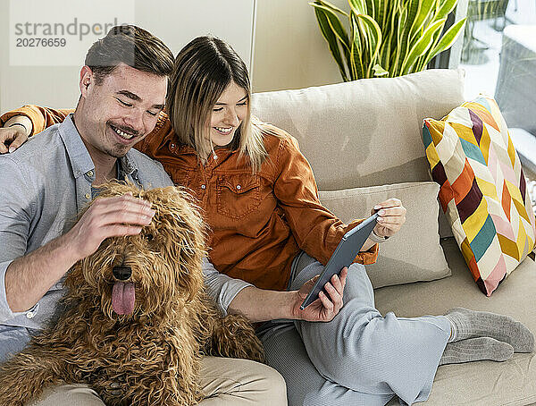 Glückliche junge Frau benutzt Tablet-PC und Mann streichelt Hund zu Hause