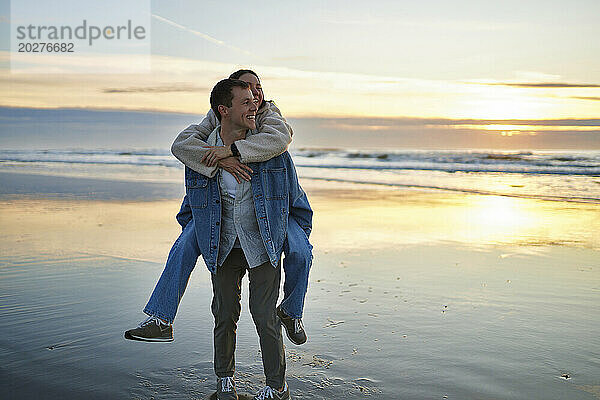 Lächelnder Freund huckepack mit Freundin am Strand bei Sonnenuntergang