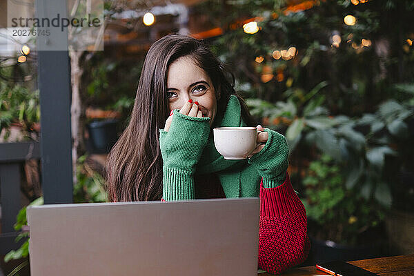 Freiberufler bedeckt Mund und hält Kaffeetasse neben Laptop im Café