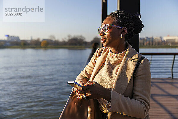 Frau mit Sonnenbrille und Smartphone in der Nähe des Flusses