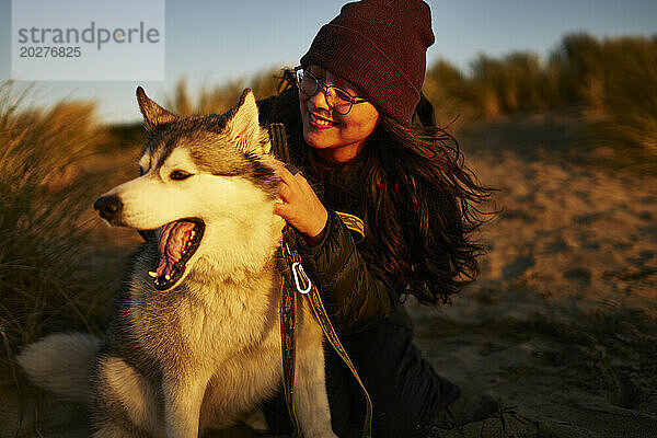 Smiling woman wearing knit hat and petting Husky dog at sunset