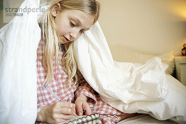 Mädchen zeichnet in Notizbuch und sitzt zu Hause im Bett