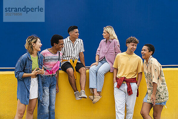 Eine Gruppe von Freunden mit farbenfroher Kleidung sitzt auf einer gelben Wand und unterhält sich