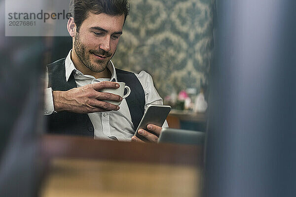 Geschäftsmann hält Kaffeetasse in der Hand und benutzt Smartphone im Café