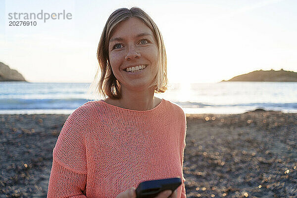 Glückliche Frau mit Mobiltelefon verbringt ihre Freizeit am Strand bei Sonnenuntergang