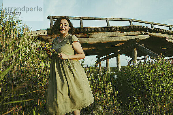 Glückliche Frau steht mit einem Strauß Wildblumen in der Nähe einer Brücke im Feld
