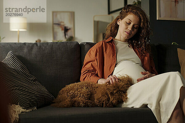 Young pregnant woman stroking dog sitting on sofa