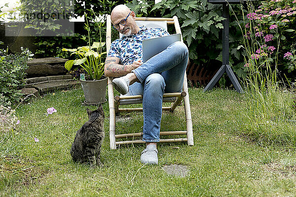 Lächelnder Freiberufler sitzt mit Laptop neben Katze im Garten