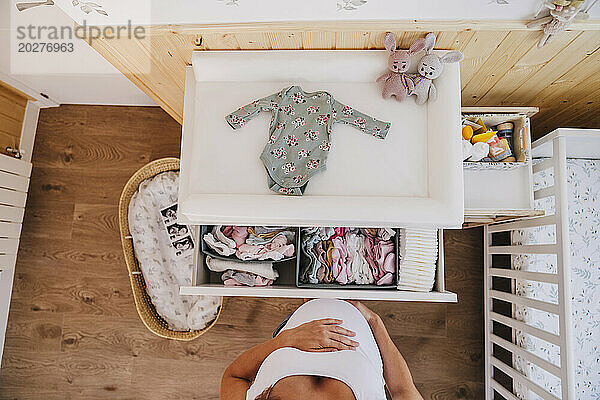 Schwangere Frau steht zu Hause neben Babykleidung