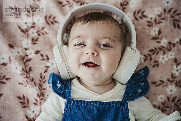Cute baby girl wearing wireless headphones lying on bed