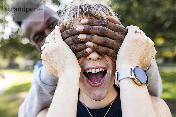 Überraschte junge Frau mit von den Händen eines Freundes bedeckten Augen im Park
