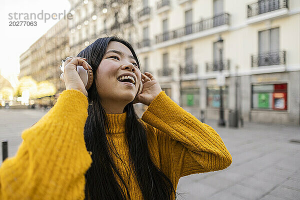 Cheerful young woman wearing wireless headphones near building