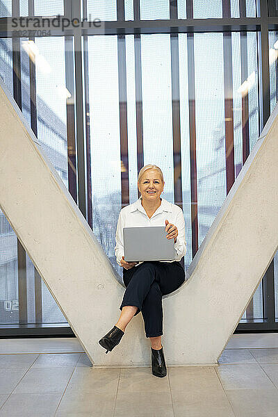 Lächelnde ältere Geschäftsfrau sitzt mit Laptop auf Säulen