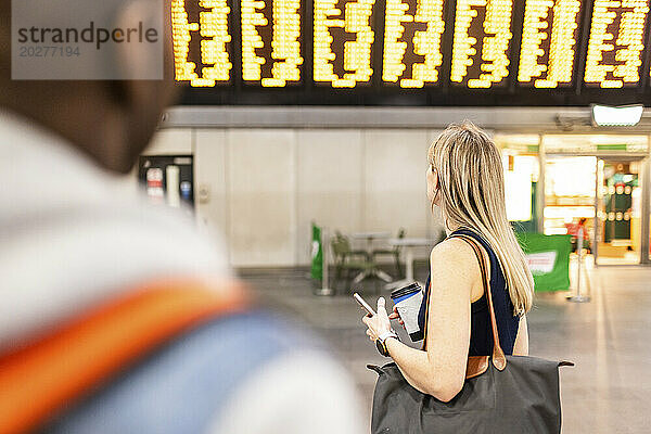 Frau trägt Tasche und liest Zugfahrplan am Bahnhof